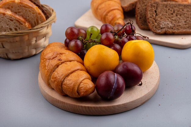 Vista laterale della frutta come uva nectacot e pluot con croissant sul tagliere e pane su sfondo grigio