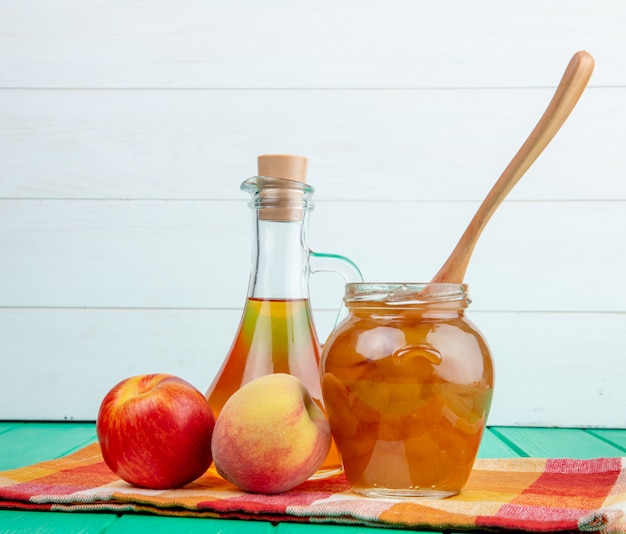 Vista laterale della frutta fresca mela con pesca e una bottiglia di olio d'oliva e marmellata di pesche in un barattolo di vetro con un cucchiaio di legno su sfondo verde in legno