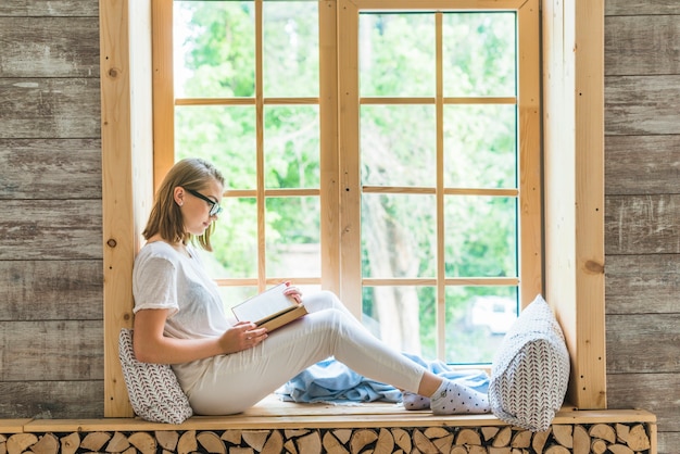 Vista laterale della giovane donna che si siede sul libro di lettura del davanzale della finestra