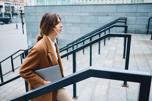 Vista laterale della giovane donna d'affari attraente in cappotto con il computer portatile che cammina con sicurezza sulla strada della città