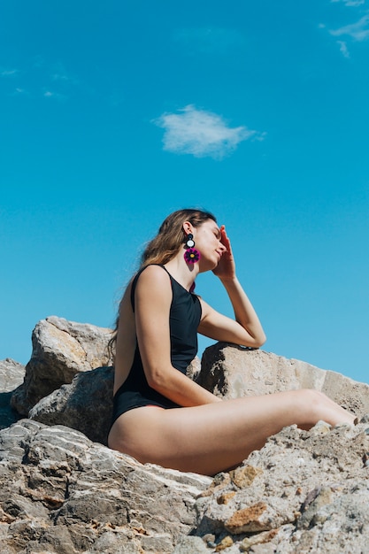 Vista laterale della giovane donna in costume da bagno nero che si siede sulla roccia