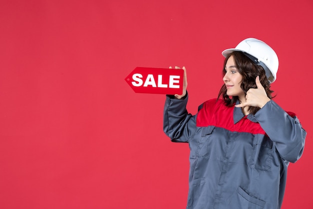 Vista laterale della lavoratrice sorridente in uniforme che indossa elmetto e indicando l'icona di vendita che fa un gesto di chiamata su sfondo rosso isolato