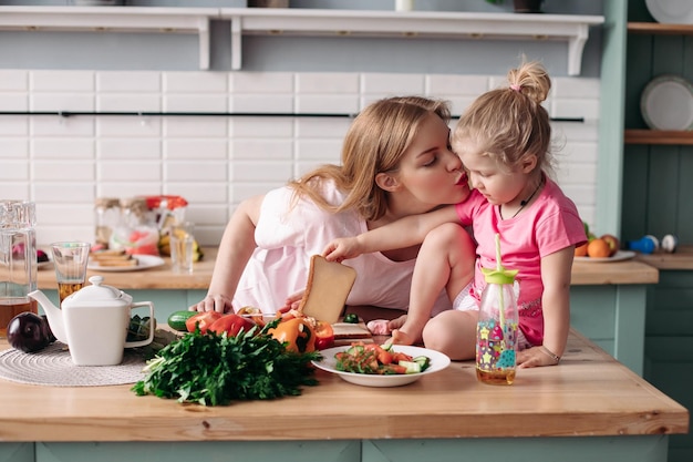 Vista laterale della madre e della figlia carine che si abbracciano e si baciano al mattino Famiglia seduta in cucina a cucinare la colazione e tagliare le verdure a casa Concetto di pranzo e cibo