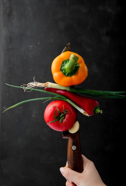 Vista laterale della mano femminile con un coltello con verdure mature fresche giallo peperone cipolla rossa peperoncino rosso e pomodoro a sfondo nero