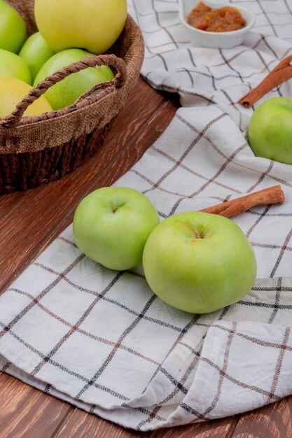 Vista laterale della merce nel carrello verde e gialla delle mele con la marmellata di mele e cannella e mele sul panno del plaid e sulla superficie di legno