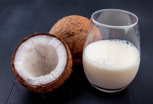 Vista laterale della noce di cocco fresca e marrone con un bicchiere di latte sulla superficie nera