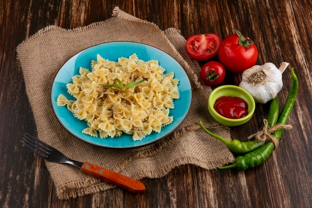 Vista laterale della pasta bollita su un tovagliolo beige piatto blu con forchetta pomodori ketchup e peperoncino su una superficie di legno