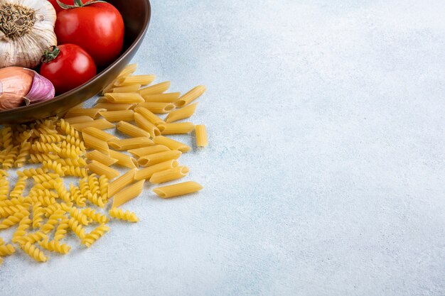 Vista laterale della pasta cruda con pomodori, aglio e cipolle in un misken contro la superficie grigia