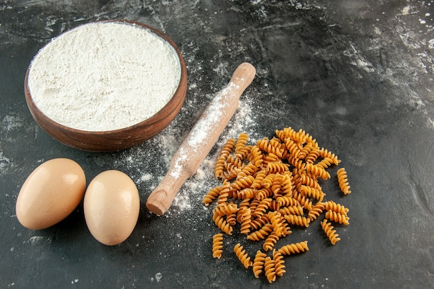 Vista laterale della pasta italiana cruda due uova e farina in una ciotola marrone mattarello su sfondo grigio