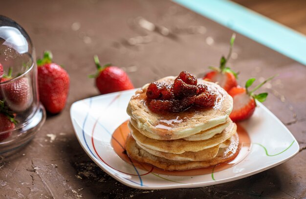 Vista laterale della pila di pancake fatti in casa con marmellata di fragole su un piatto e fragole fresche su rustico