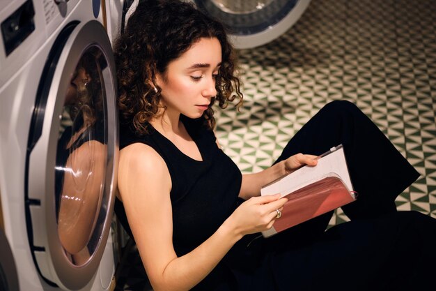 Vista laterale della ragazza attraente che legge sognante il libro in attesa di lavaggio nella moderna lavanderia self-service