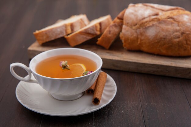 Vista laterale della tazza di hot toddy con fiore di limone all'interno e cannella sul piattino con taglio e fette di pane croccante sul tagliere su sfondo di legno