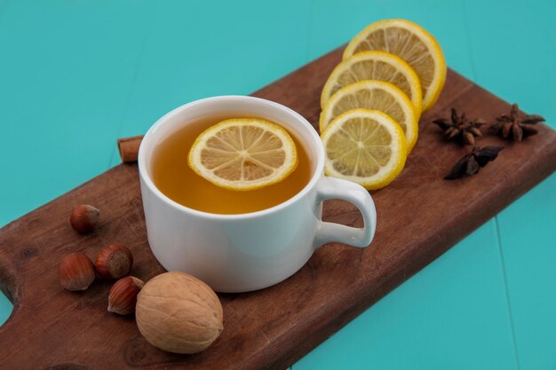 Vista laterale della tazza di tè con fette di limone e cannella con noci noce sul tagliere su sfondo blu