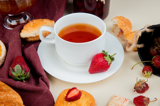 Vista laterale della tazza di tè con il cioccolato dell'inceppamento del bigné dei rotoli delle fragole sulla tavola bianca