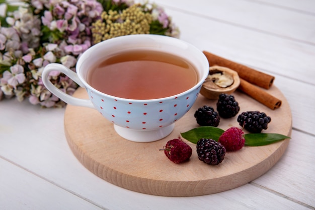 Vista laterale della tazza di tè con lamponi noci cannella e more su un tagliere con fiori su una superficie bianca