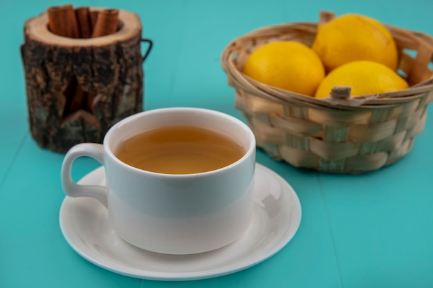 Vista laterale della tazza di tè con una ciotola di cannella e un cesto di limoni su sfondo blu