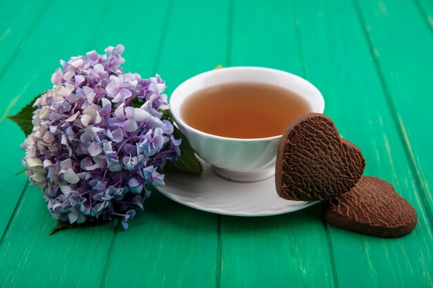 Vista laterale della tazza di tè e biscotti a forma di cuore con fiori su sfondo verde