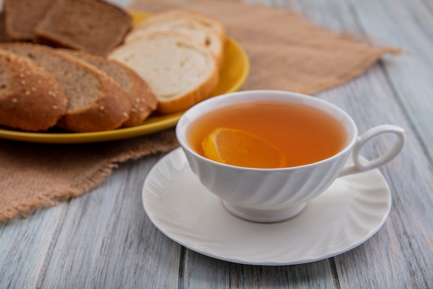 Vista laterale della tazza di toddy caldo sul piattino con pane a fette nel piatto su tela di sacco su sfondo di legno