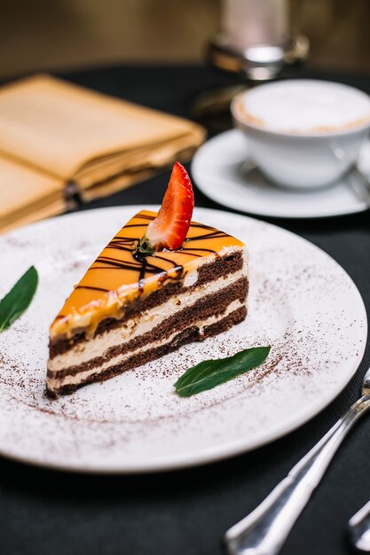 Vista laterale della torta al cioccolato a strati ricoperta di caramello una fetta di fragola su un piatto bianco