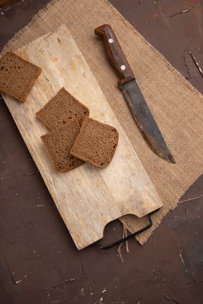 Vista laterale delle fette del pane di segale sul tagliere e coltello su tela di sacco su fondo marrone rossiccio con lo spazio della copia