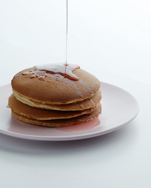 Vista laterale delle frittelle in piastra e sciroppo di marmellata di fragole che si versano su sfondo bianco