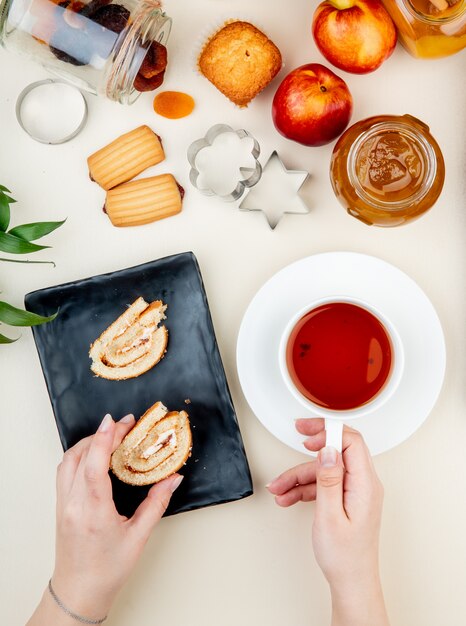 Vista laterale delle mani della donna che tengono la fetta del rotolo e la tazza di tè con il barattolo delle pesche dei biscotti delle marmellate dell'uva passa su bianco