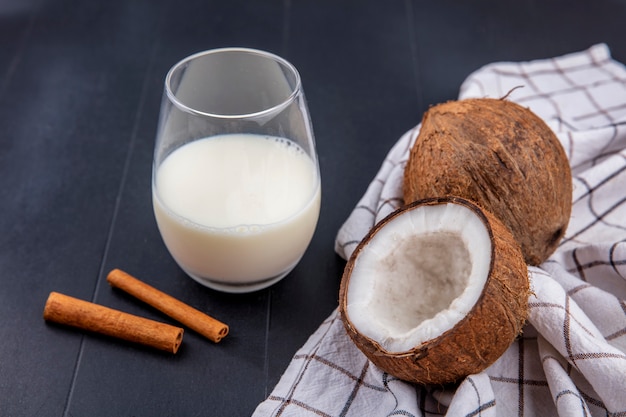 Vista laterale delle noci di cocco con un bicchiere di latte con i bastoncini di cannella su una tovaglia controllata su una superficie di legno