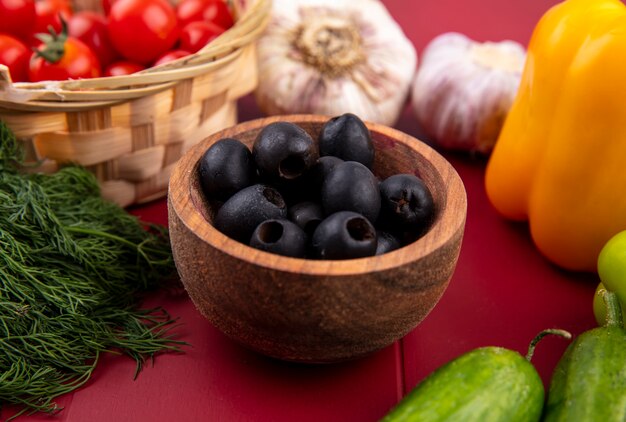 Vista laterale delle olive nere in ciotola con il cetriolo del pepe dell'aneto dell'aneto del pomodoro su superficie rossa