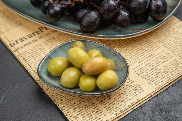 Vista laterale delle olive verdi organiche fresche e dei fasci di uva nera su un vecchio giornale su un fondo scuro