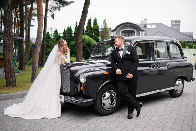 Vista laterale dello sposo elegante e bello in smoking nero che si appoggia a un'auto retrò e guarda alla sposa che sta di fronte e lo guarda adorabile durante la passeggiata di nozze all'aperto