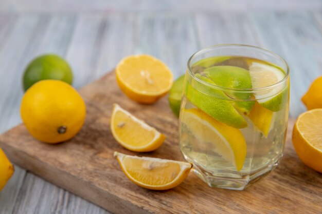 Vista laterale detox acqua in un bicchiere con spicchi di lime e mezza arancia e limone su un tagliere