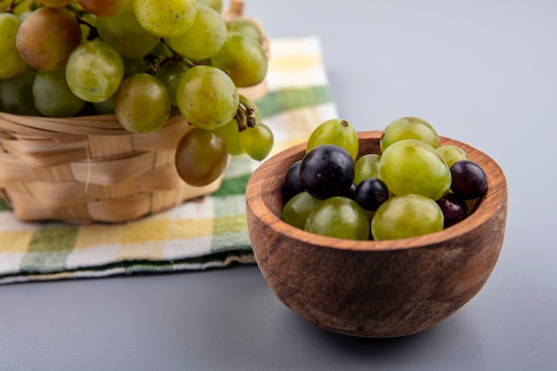 Vista laterale di acini d'uva in una ciotola con cesto di uva sul panno plaid e sfondo grigio