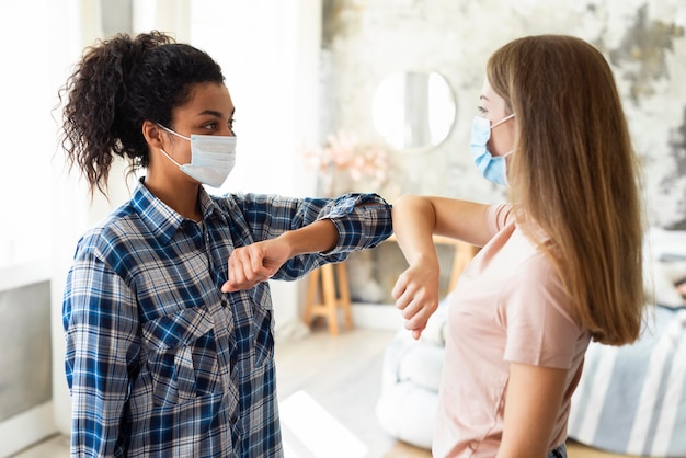 Vista laterale di amiche con maschere mediche che praticano il saluto di gomito