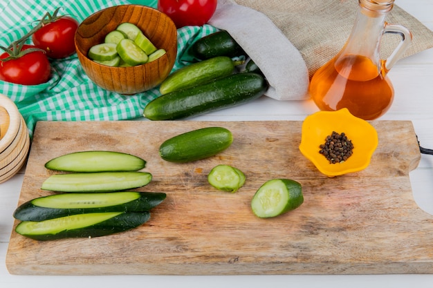 Vista laterale di cetrioli interi e tagliati e tagliati e pepe nero sul tagliere con pomodori olio fuso ciotola di fette di cetrioli e cetrioli fuoriuscita dal sacco su legno