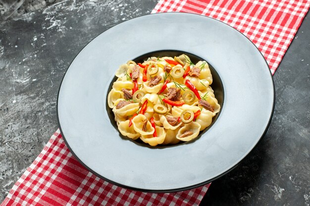 Vista laterale di deliziose conchiglie con verdure su un piatto e coltello su asciugamano spogliato rosso su sfondo grigio