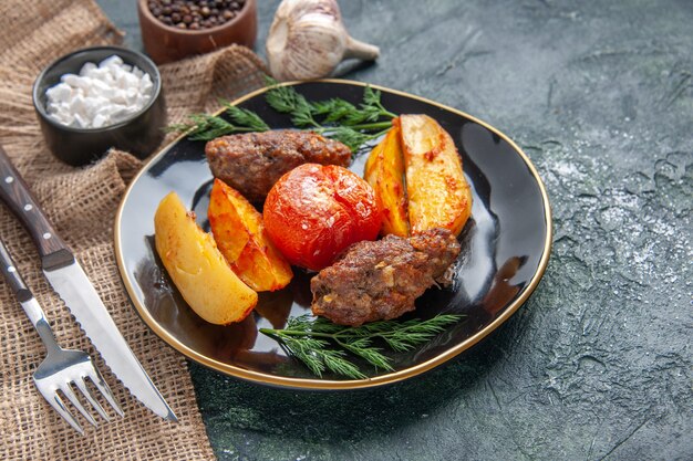 Vista laterale di deliziose cotolette di carne al forno con patate e pomodori su un piatto nero spezie aglio posate su sfondo nero verde colori mix