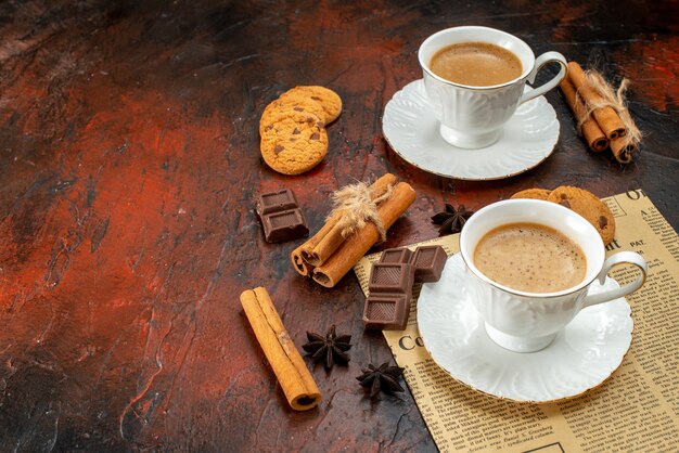 Vista laterale di due tazze di caffè biscotti alla cannella lime barrette di cioccolato su un vecchio giornale sul lato sinistro su superficie scura