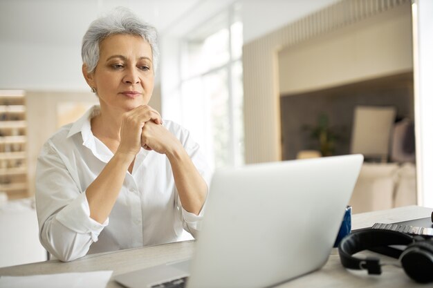 Vista laterale di felice di mezza età imprenditrice con corti capelli grigi che lavora al computer portatile nel suo elegante ufficio con le mani sulla tastiera, digitando la lettera, condividendo buone notizie