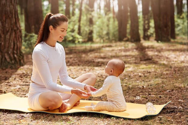 Vista laterale di felice giovane madre sportiva seduta su karemat nella foresta con il suo bambino neonato