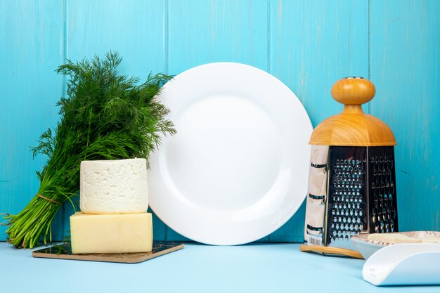 Vista laterale di feta bianco con aneto e grattugia e piatto bianco vuoto sul blu