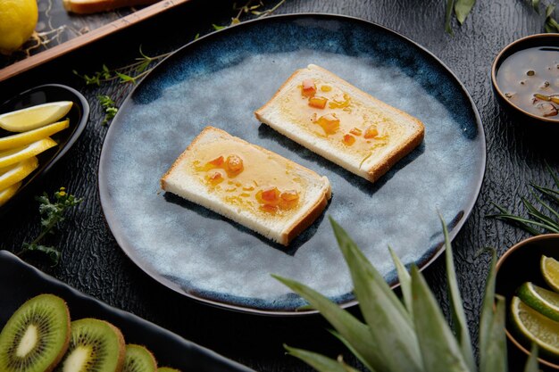 Vista laterale di fette di pane spalmate di marmellata con agrumi intorno come ananas kiwi arancia lime fette di limone e mandarino con fette di pane al limone in vassoio su sfondo nero