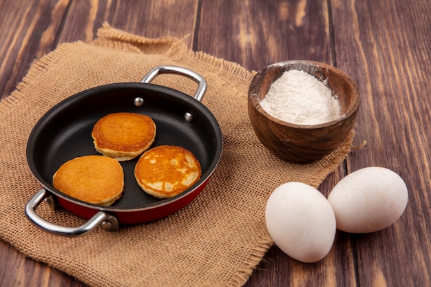 Vista laterale di frittelle in padella e ciotola di farina su tela di sacco con uova su sfondo di legno