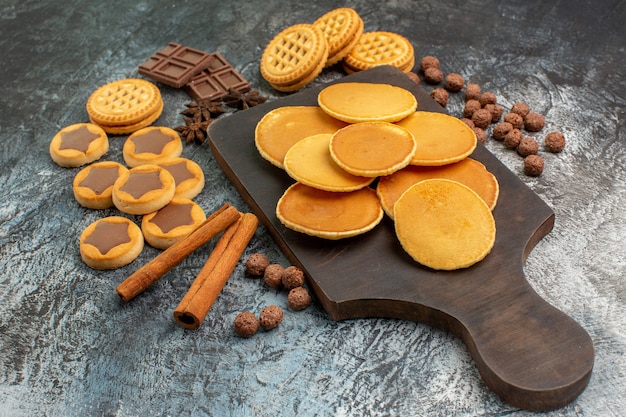 Vista laterale di frittelle su piatto di legno con biscotti e dolci su fondo grigio