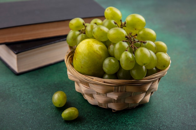 Vista laterale di frutti come uva e pluot nel cesto con libri chiusi su sfondo verde