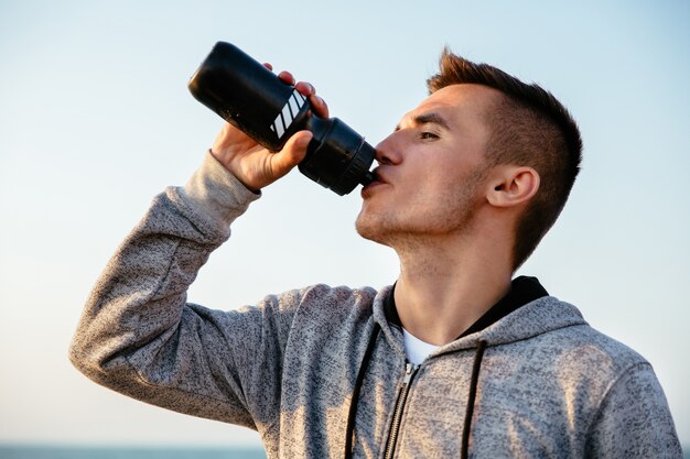 Vista laterale di giovane sportivo che beve un acqua dalla bottiglia dopo l&#39;esecuzione, allenamento