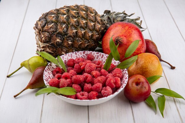 Vista laterale di lamponi in una ciotola con melograno ananas pesca prugna e mela intorno su bianco