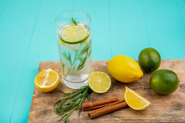 Vista laterale di limoni colorati con rinfrescante acqua estiva in un bicchiere su una tavola di cucina in legno con bastoncini di cannella sulla superficie blu