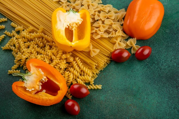 Vista laterale di pasta cruda con spaghetti crudi e peperoni colorati e pomodorini su una superficie verde