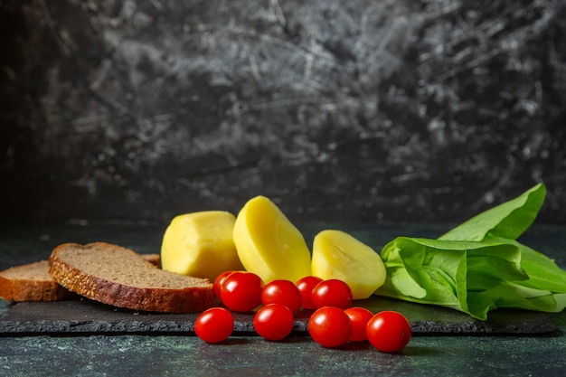 Vista laterale di patate fresche tagliate e fette di pane dietetico pomodori fascio verde sul tagliere di legno su sfondo di colori mescolare nero verde con spazio libero