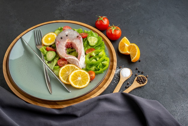 Vista laterale di pesce crudo e verdure fresche fette di limone e posate impostato su un piatto grigio spezie asciugamano di colore scuro sulla superficie nera